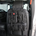 A black tactical vehicle seat cover with multiple zippered compartments, MOLLE webbing, and a red cross patch, shown installed on a car seat inside a vehicle.