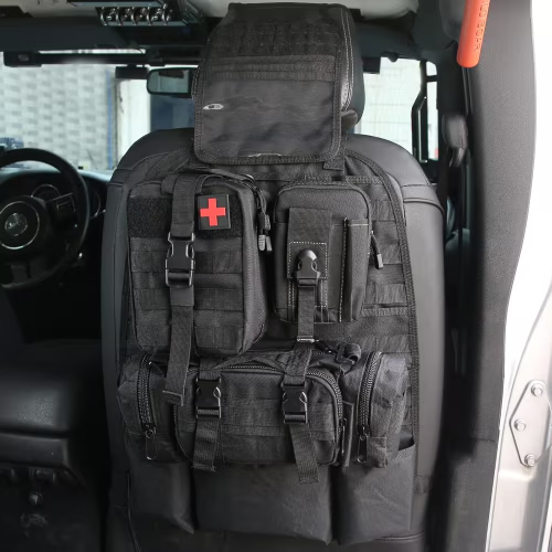 A black tactical vehicle seat cover with multiple zippered compartments, MOLLE webbing, and a red cross patch, shown installed on a car seat inside a vehicle.