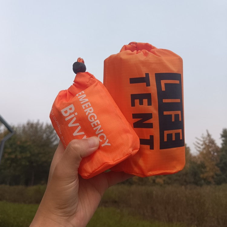 Two orange emergency survival bags labeled 'Bivvy' and 'Life Tent' held in a hand with a blurred natural background.