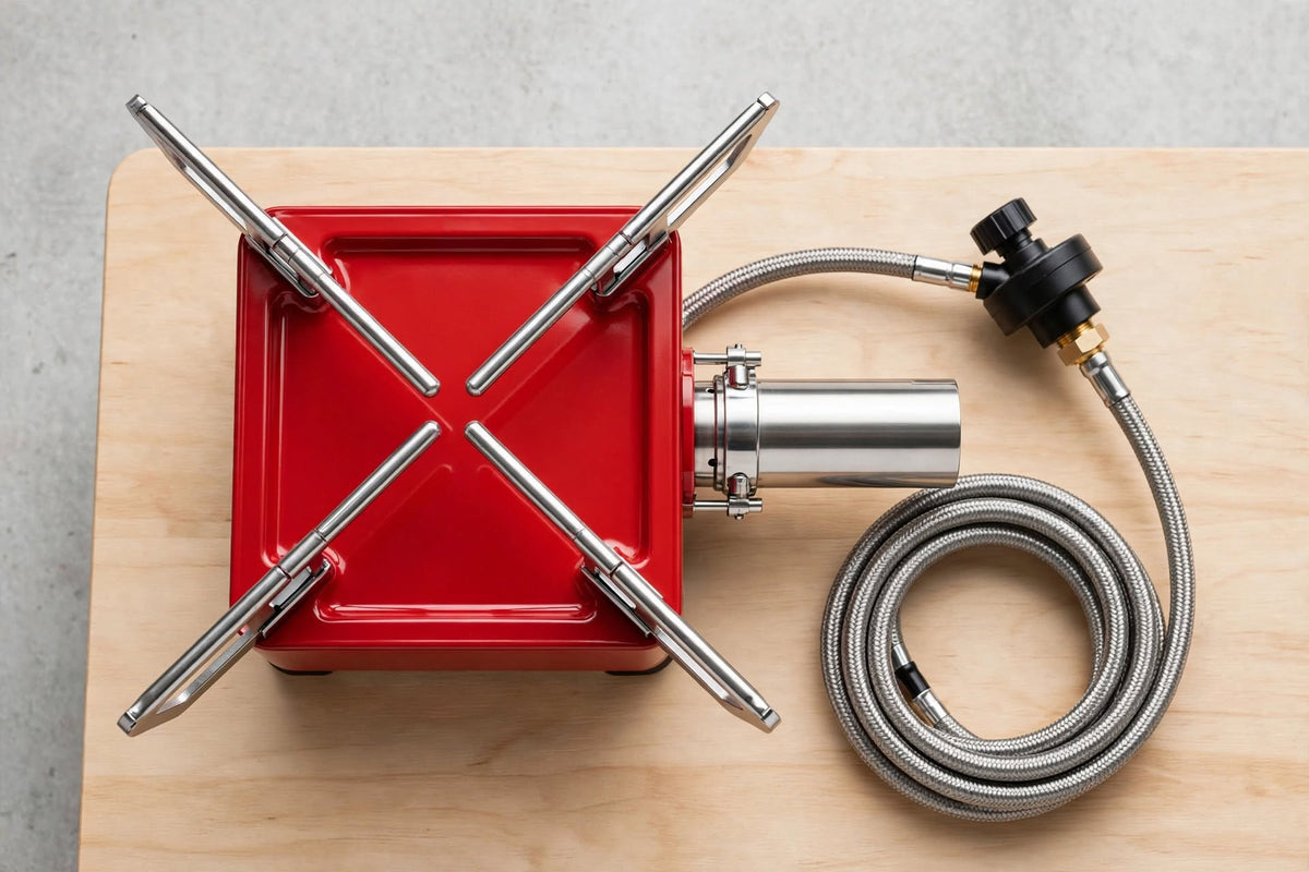 A red portable gas stove top with four metallic support legs and a braided metal gas hose connected to a black and brass regulator, displayed on a light wood board against a gray background.