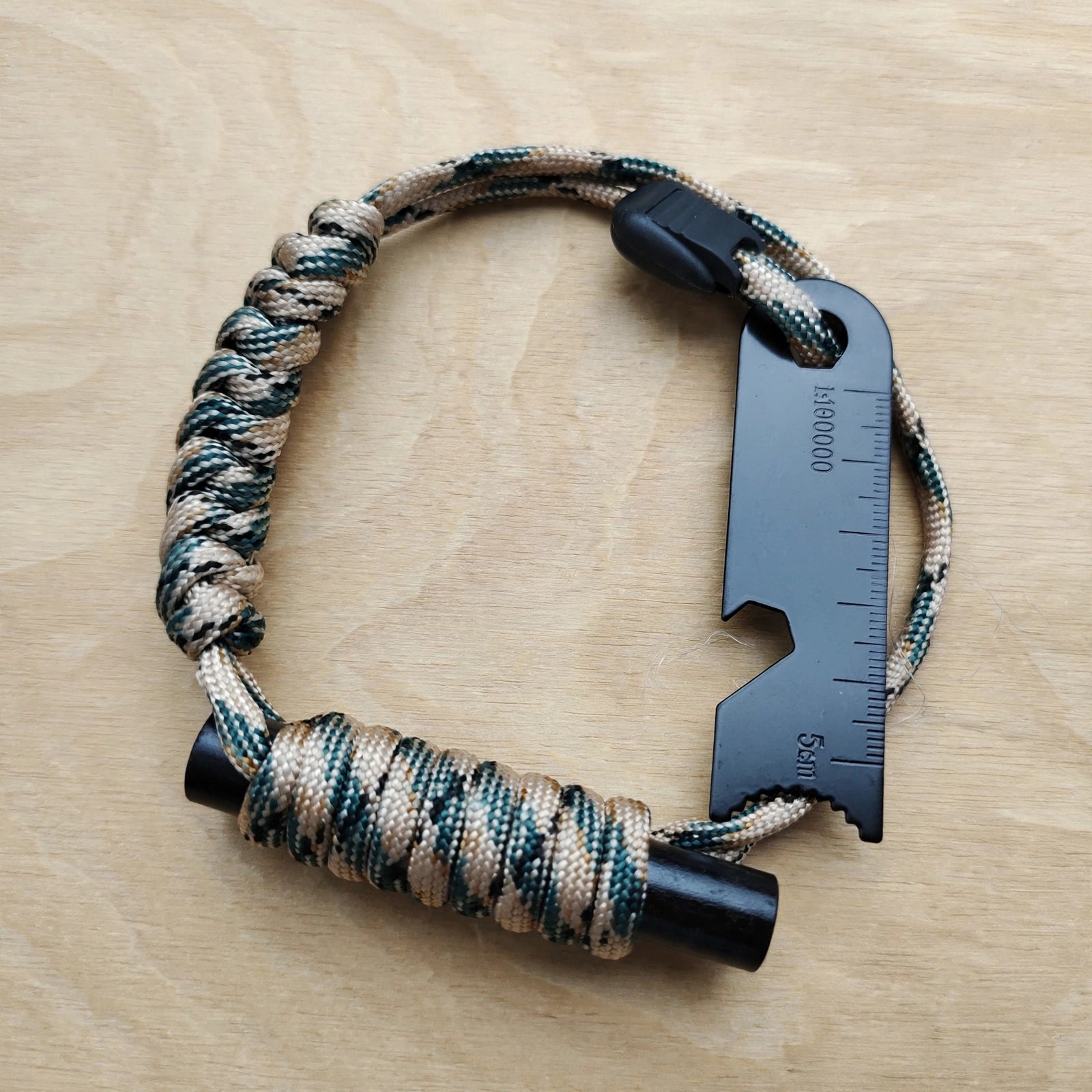 A tactical paracord bracelet with a black multi-tool keychain, shown on a light wood background. The paracord is woven in a camouflage pattern of beige, green, and black, and features a black plastic buckle and a black multi-tool keychain with a ruler scale.