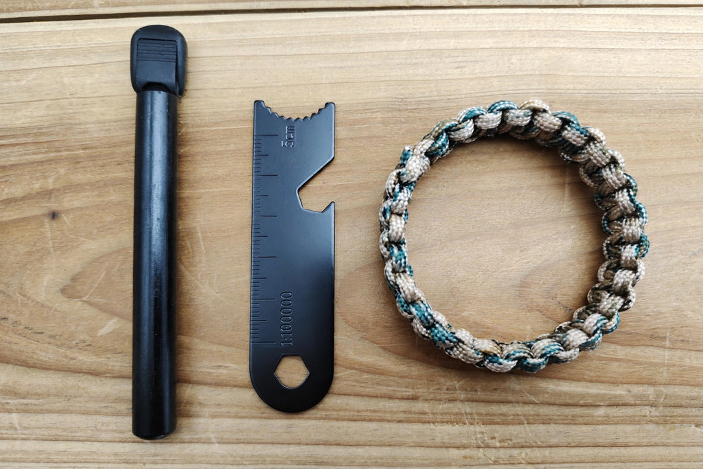 A handmade paracord bracelet in beige, brown, and teal woven alongside a black fire starter tool and a black metal divider tool on a light wood background.