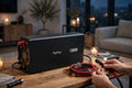 A black 1300W portable power station sits on a wooden table in a cozy, dimly lit living room. A person's hands are shown holding red and black cables connected to the power station, with a flashlight visible.