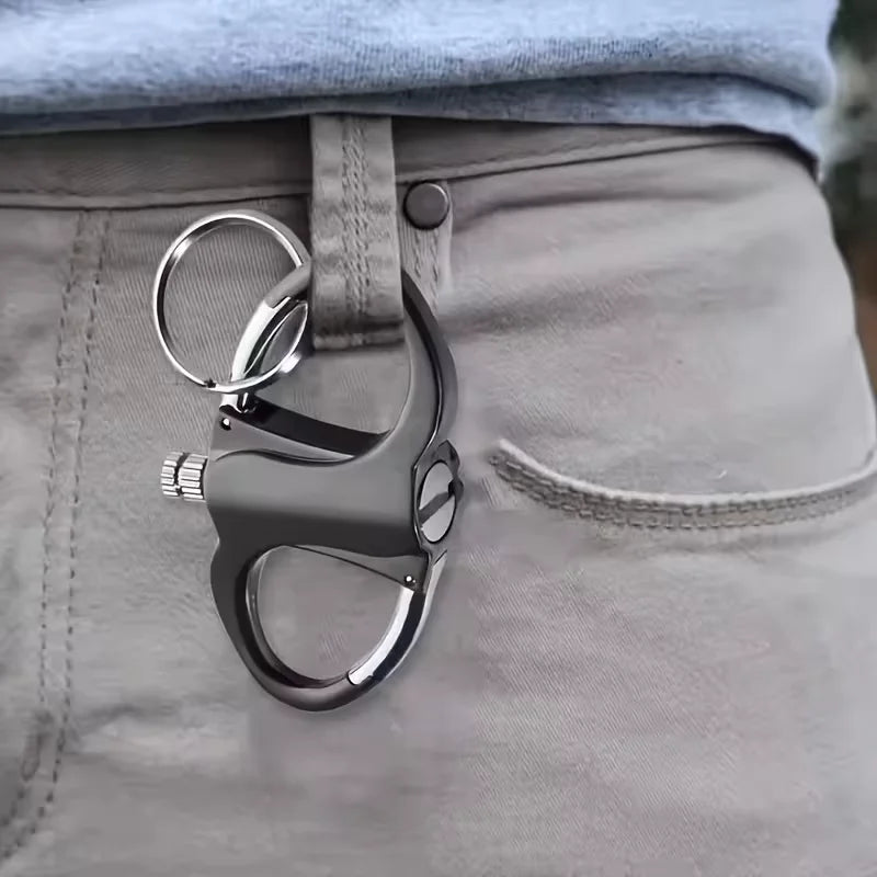 A close-up of a gray and silver self-defense keychain, specifically a pepper spray trigger with a ring, clipped to the waist belt of a person wearing light gray khaki pants.