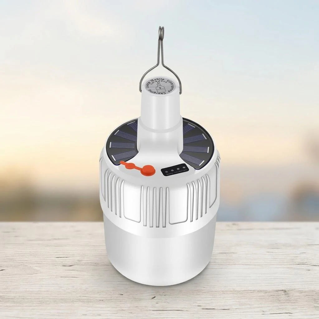 A white cylindrical portable lantern with a solar panel top, hanging loop, and small orange accent, shown on a light wooden surface against a softly blurred outdoor background.