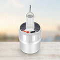 A white cylindrical portable lantern with a solar panel top, hanging loop, and small orange accent, shown on a light wooden surface against a softly blurred outdoor background.