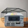 A silver metal power supply unit with a black front panel, featuring a USB port, AC220V 50Hz input, and a green power indicator light. It is shown on a wooden surface with a black metal mesh rack behind it.
