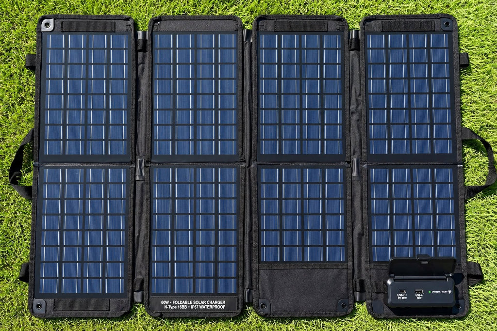 A portable foldable solar panel pack laid flat on green grass. The black fabric case is unfolded into six panels, each covered with blue solar cells, and a small black power management module is attached at the lower right.