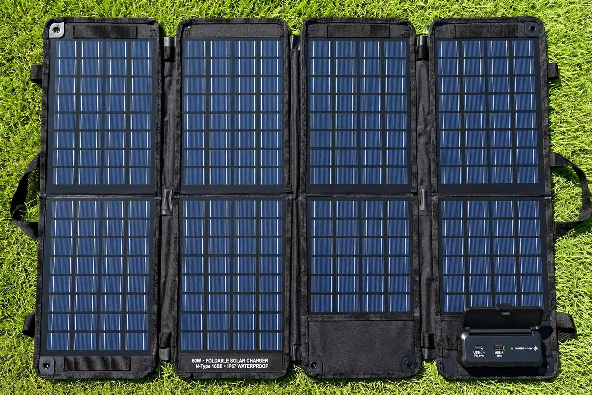 A portable foldable solar panel pack laid flat on green grass. The black fabric case is unfolded into six panels, each covered with blue solar cells, and a small black power management module is attached at the lower right.